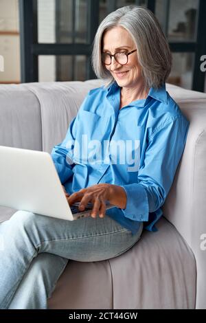 Happy 60s mature woman using laptop computer sitting on couch at home. Stock Photo