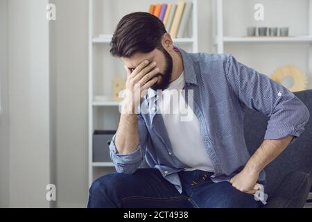 Sad man sitting at home, feeling bad and touching face with hand Stock Photo