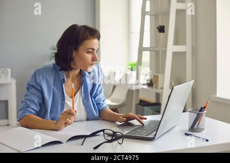 Woman talking to somebody and making notes during online conversation on laptop at home Stock Photo