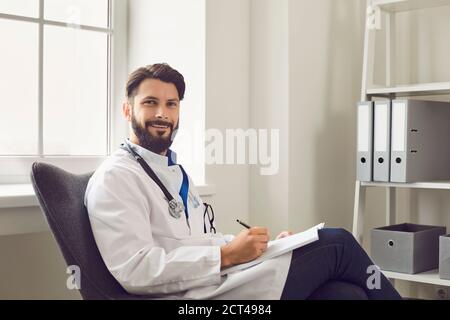 Handsome young medical doctor in lab coat sitting at hospital. Medical worker writing prescription form at office Stock Photo