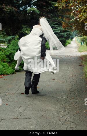 groom running away with bride on his back outdoors in park alley Stock Photo