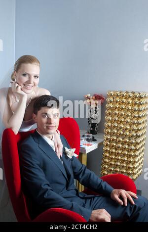 Groom sitting in red chair and smiling bride behind with empty wall for copy space Stock Photo
