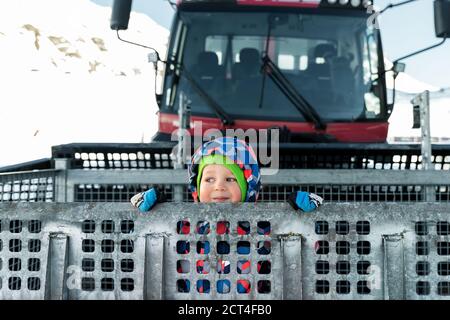 Cute adorable playful happy toddler boy inside red modern snowcat ratrack snowplow box grooming standing on peak alpine skiing resort Ischgl Austria Stock Photo
