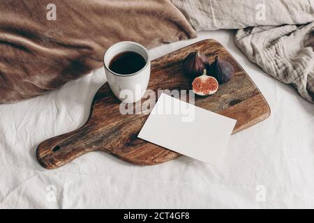 Autumn breakfast in bed composition. Blank greeting card mockup, cup of coffee and fig fruit on wooden cutting board. White linen background. Velvet c Stock Photo