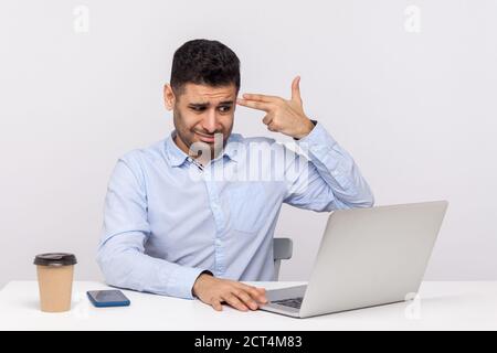Depressed male employee sitting office working on laptop, pointing finger gun gesture to head, committing suicide from stressful job, tired of overwor Stock Photo