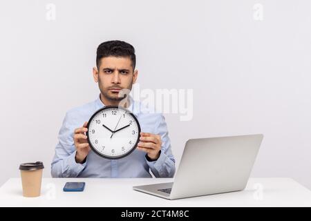 Time management. Serious displeased man employee sitting in office workplace with laptop, holding big clock and looking at camera with strict angry ex Stock Photo