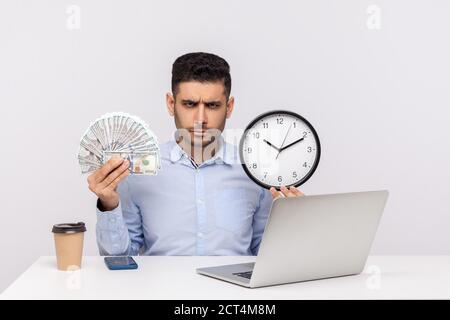 Time is money! Bossy angry businessman sitting in office workplace, holding big clock and dollar banknotes, frowning looking at camera strict expressi Stock Photo