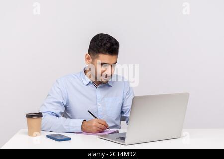 Busy man employee working on laptop and writing paper notebook, preparing report doing paperwork, journalist taking notes in his office workplace. ind Stock Photo