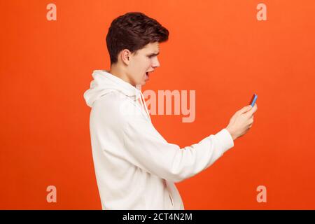 Profile portrait of shocked teenager in white casual sweatshirt looking at screen of mobile phone with excited face, receiving email with sale coupons Stock Photo