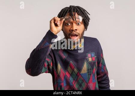 Extremely shocked wondered african man with dreadlocks looking at camera with widely open mouth and big eyes, putting off glasses, surprised. Indoor s Stock Photo