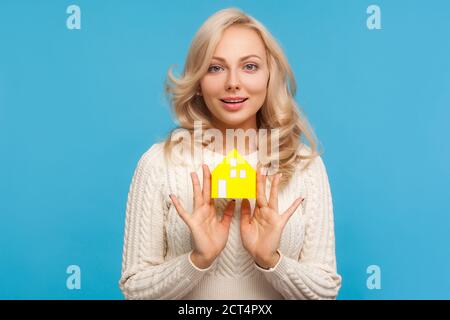 Attractive blond woman with curly hair showing paper house at camera, real estate agency, accommodation rent. Indoor studio shot isolated on blue back Stock Photo