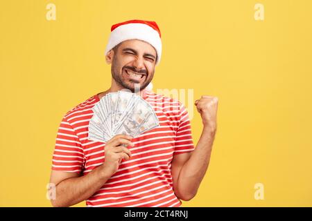 Extremely happy pleased bearded man in red t-shirt and santa claus hat showing yes gesture holding fan of dollars, perks and bonuses, success. Indoor Stock Photo