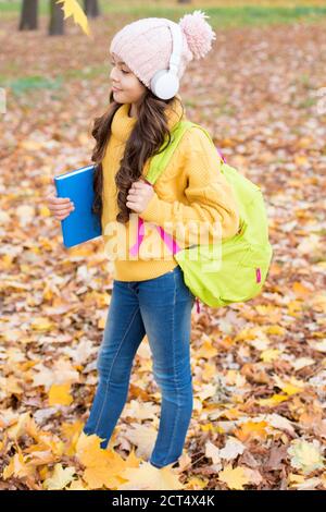 knowledge day. kid fall clothes fashion. happy kid walk in autumn park. small girl wear headphones. back to school. modern education. child with backpack and book in forest. Waiting for inspiration. Stock Photo