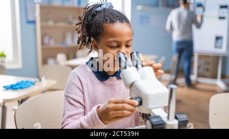 Portrait of Smart Little Schoolgirl Looking Under the Microscope. In Elementary School Classroom Cute Girl Uses Microscope. STEM science, technology Stock Photo