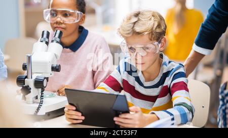 Elementary School Science Classroom: Boy Uses Digital Tablet Computer to Check Information on the Internet while Enthusiastic Cute Little Girl Uses Stock Photo