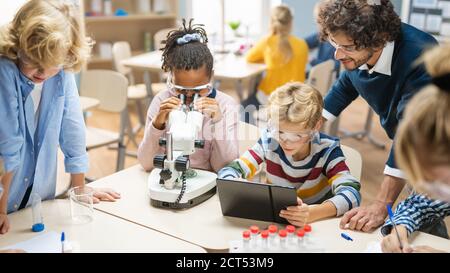 Elementary School Science Classroom: Cute Little Girl Looks Under Microscope, Boy Uses Digital Tablet Computer to Check Information on the Internet Stock Photo