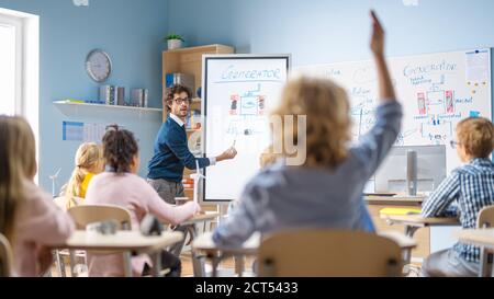 Brilliant Schoolboy Listens Attentively to His Teacher Explaning Lesson and Raises His Hand with a Question. In Elementary School with Group of Bright Stock Photo
