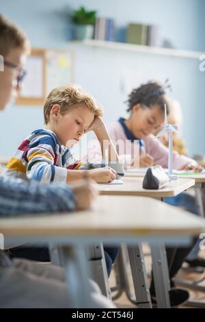 In Elementary School Classroom Brilliant Caucasian Boy Writes in Exercise Notebook, Taking Test and Writing Exam. Junior Classroom with Group of Stock Photo