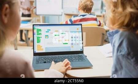 Elementary School Science Class: Over the Shoulder Little Boy and Girl Use Laptop with Screen Showing Programming Software. Physics Teacher Explains Stock Photo