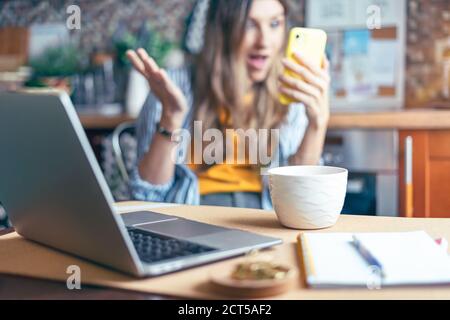 Business woman having a facetime video call. Surprised girl working from home office kithcen and drinking coffee. Using computer and looking at cellph Stock Photo