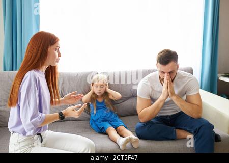 sad, desperate little girl during parents quarrel, she clog the ears sitting on sofa at home, angry parents fighting. worried upset small daughter hur Stock Photo