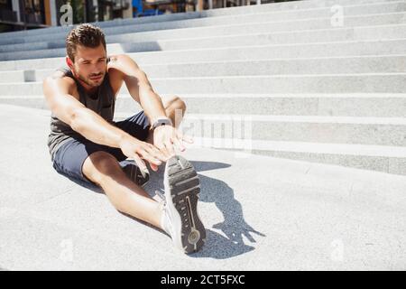 Young man exercising outside. A view of sportsman sitting beside steps and stretching arms and leg. Doing yoga exercise. Training and having workout Stock Photo