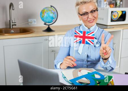 cool modern english teacher hold online lesson via webcam on laptop, she looks at screen smiling, britain flag in hands Stock Photo
