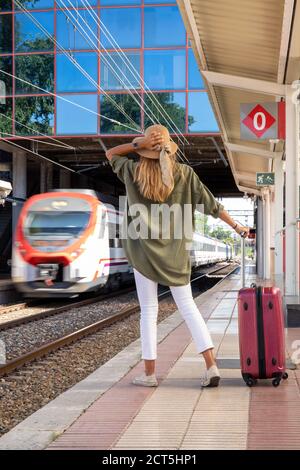 Back view of anonymous female traveler with suitcase standing on platform and waiting for train on sunny day on railway station Stock Photo