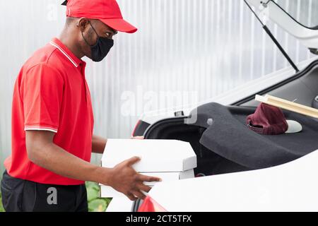 side view on young black courier in mask putting order in boxes at car, during coronavirus quarantine Stock Photo