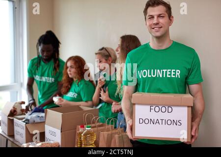 portrait of affable volunteer man in green t-shirt holding donation box in hands while his friends collecting clothes and food in the background Stock Photo