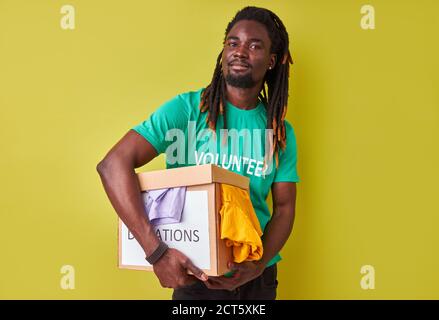 portrait of black afro volunteer man with box full of clothes, young male with dreadlocks engaged in charity, love helping others Stock Photo
