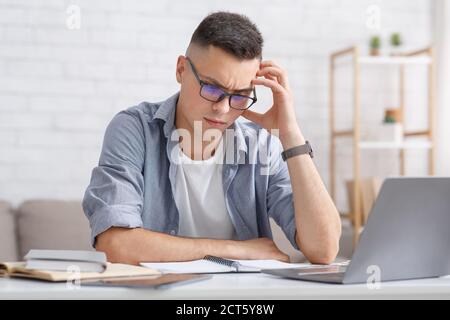 Difficult task and home remote education. Brainstorming of young guy sitting at table with laptop and looking at notebook in interior of living room Stock Photo