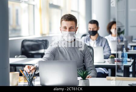 Attractive worker at workplace takes pencil in his hands behind laptop. Guy in protective mask sits at table in office interior during quarantine Stock Photo
