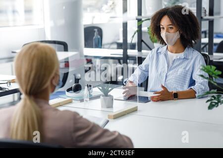 Young business workers in protective mask talking to each other in office through partition to prevent COVID-19 infection Stock Photo