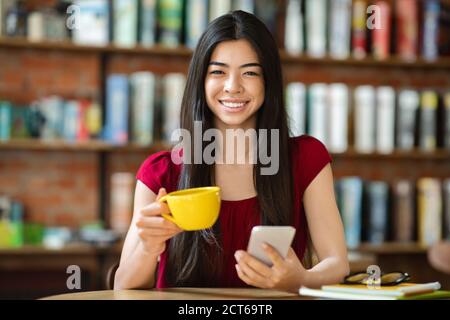 Coffee Break. Asian Woman Relaxing With Smartphone And Hot Drink In Cafe Stock Photo