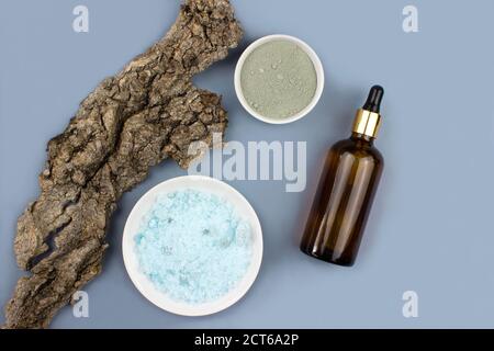 Sea salt, serum in brown dropper glass, clay mask and tree bark flat lay on blue background top view. Organic, natural cosmetic. Skincare, spa.