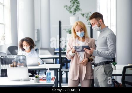 Colleagues in protective masks discuss project and look at tablet in coworking office Stock Photo