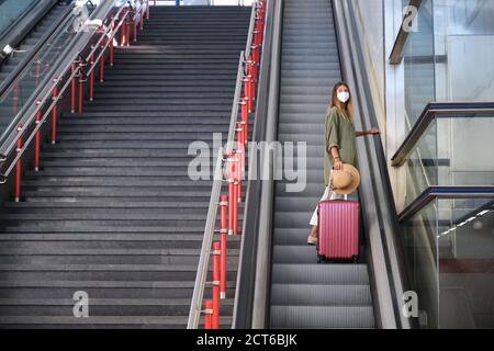 Young woman wearing face mask, and carrying a suitcase and a hat on the escalators. Travel concept. Stock Photo