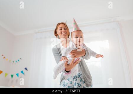 A smiling mother holds a cute baby in her arms, playing with it, imitating the flight of an airplane. Holiday hats on their heads. Concept of motherho Stock Photo