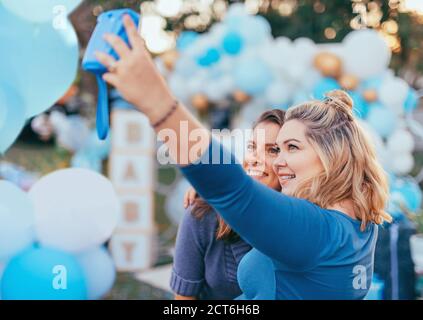 Female friends taking selfie with pregnant woman at a baby shower. Mobile photography, party decorations in white and blue colors, baby boy Stock Photo