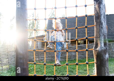 Cute caucasian blond baby girl plays at background, climbing up and down the net Stock Photo