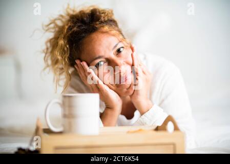 Cheerful beautiful young adult caucasian woman portrait lay down in the bedroom on the bed with cup of tea or coffee - happy peole at home enjoy the m