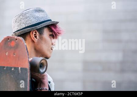 Portrait of handsome young teenager man boy looking in front of him and holding a skateboard - diversity concept and alternative violet hair and hat p Stock Photo