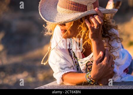 Portait of beautiful young trendy woman with cowboy hat and accessories like bracelets lay down on a wall bench enjoying the sunset light - people in Stock Photo