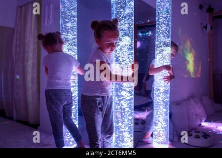 Child in therapy sensory stimulating room, snoezelen. Autistic child interacting with colored lights bubble tube lamp during therapy session. Stock Photo