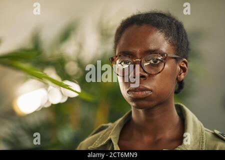 Head and shoulders portrait of contemporary African-American woman wearing glasses and looking at camera while standing in modern office with plants, copy space Stock Photo