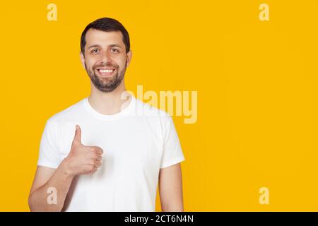 Young man with a beard in a white t-shirt making a happy hand gesture. Confirming facial expression showing success while looking at the camera. Stands on isolated yellow background Stock Photo