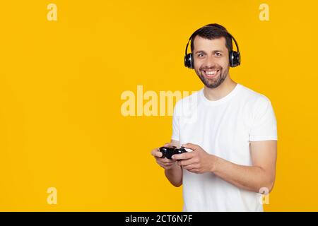Young man with beard in white t-shirt gamer playing video games with headphones smiling and looking at the camera. Stands on isolated yellow background. Stock Photo