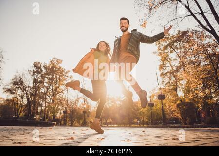 Low angle view full length photo of cheerful two young people girl guy hold hand jump in autumn october town park