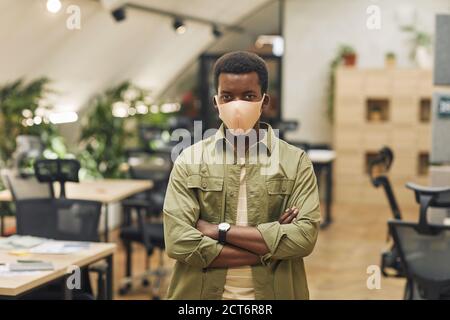 Waist up portrait of confident African-American man wearing mask in office and looking at camera while standing with arms crossed in office, copy space Stock Photo